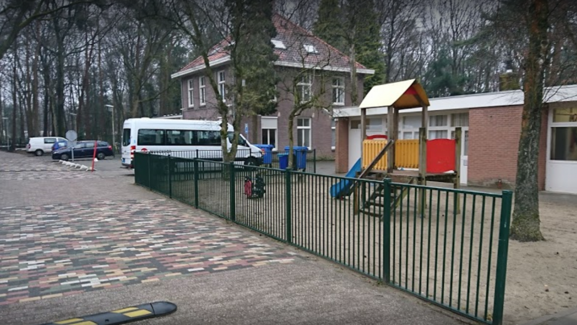 buitenterrein met rechts een gebouw met plat dak en daarvoor een kleurig speeltoestel met rondom een hek om het plein daarachter een gebouw met drie verdiepingen en puntdak en daarvoor geparkeerde auto's en een wit personenbusje en veel bomen op en rond het terrein