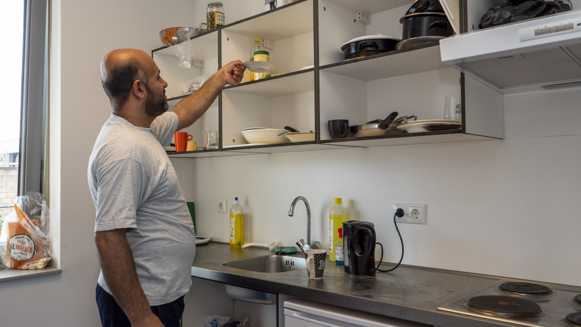 bewoner pakt iets van een plank in een keuken met waterkoker, koelkast en kookplaat