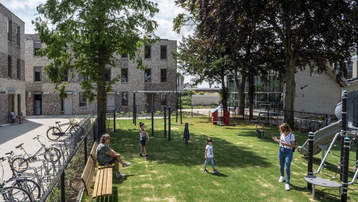 grasveld met speeltoestellen en bankjes met bewoners en spelende kinderen daaromheen fietsenrekken met fiesten en gebouwen