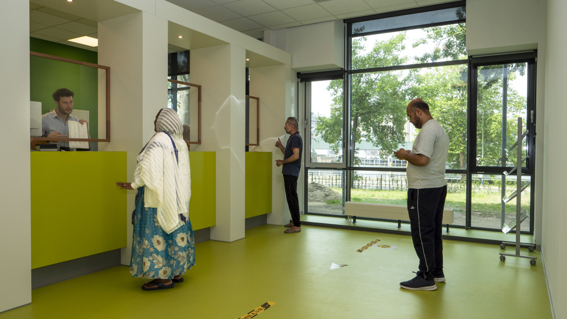 Informatiebalie met grote ramen, witte en groene muren, een groene vloer, bewoners met vragen en achter de balie medewerkers die hen helpen