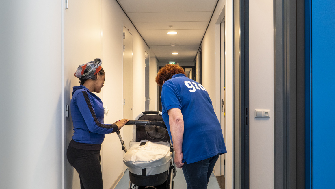 bewoonster met baby in kinderwagen op de gang bij gezondheidszorg asielzoekers, medewerkster kijkt in kinderwagen