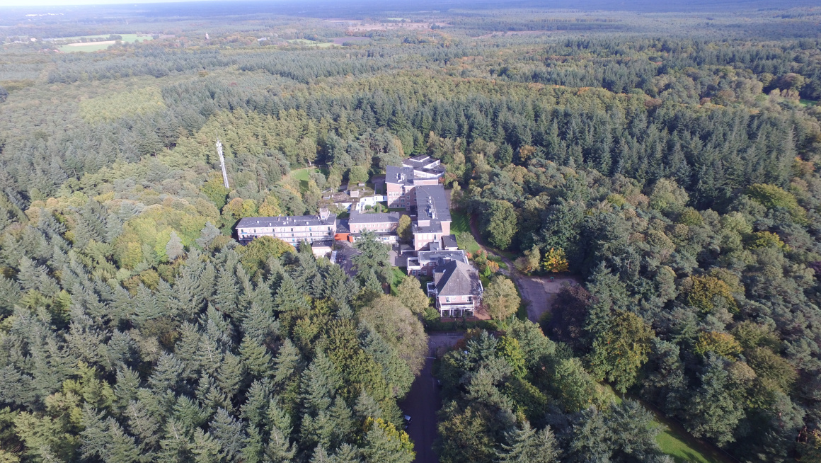 Luchtfoto azc-gebouwen met grijze daken met rondom veel bomen