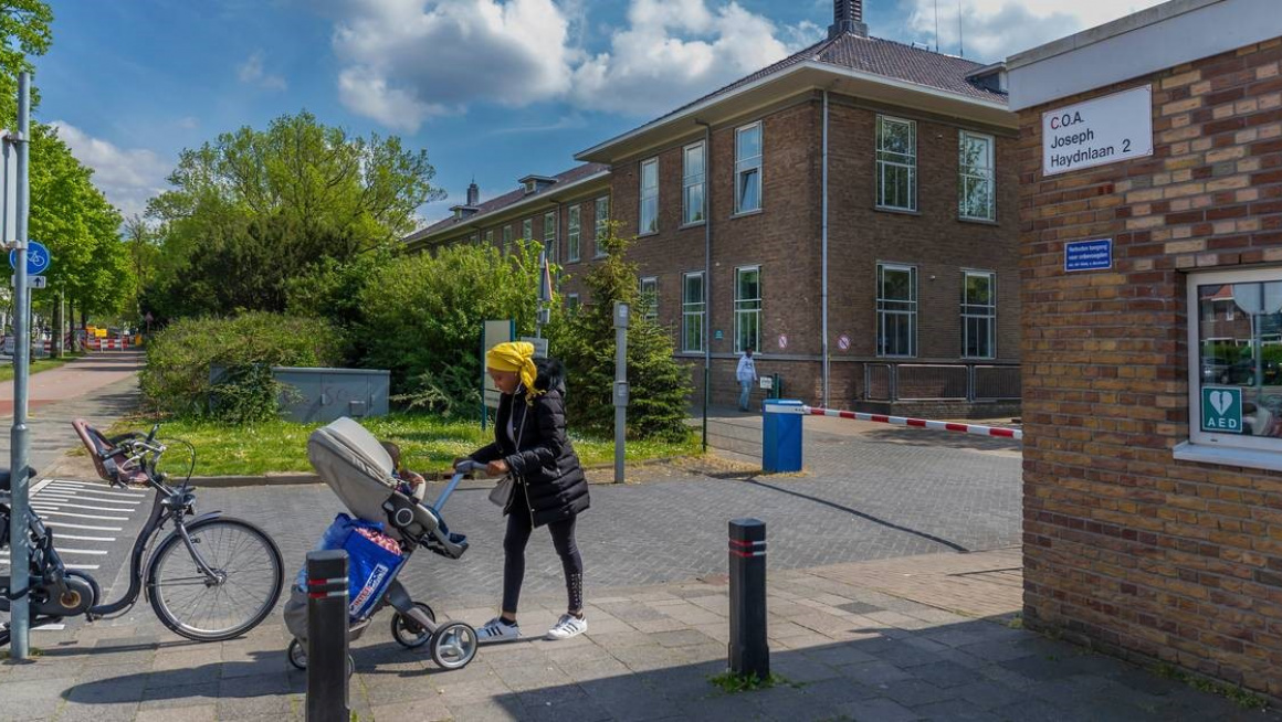 entree azc met rechts een gebouw met receptie en daar tegenover een woongebouw met daarnaast struiken en bomen op de voorgrond een vrouw met gele hoofddoek en een kinderwagen