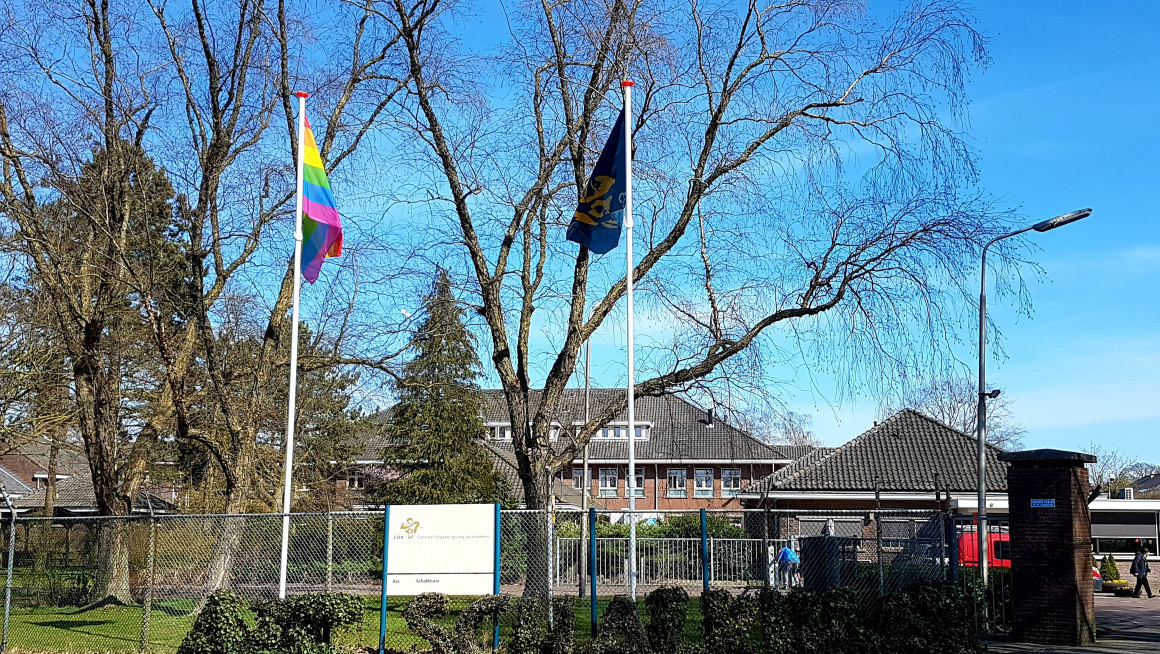 Ingang azc met op de achtergrond gebouwen, daarvoor bomen, daarvoor struiken met een bord met COA en azc Schalkhaar erop en daarchter twee vlaggenmasten met de COA-vlag en de lhbti-vlag