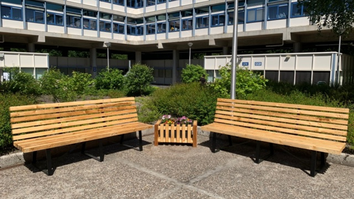 Gebouw met daarvoor begroeiing en twee bankjes met bloembak ertussen