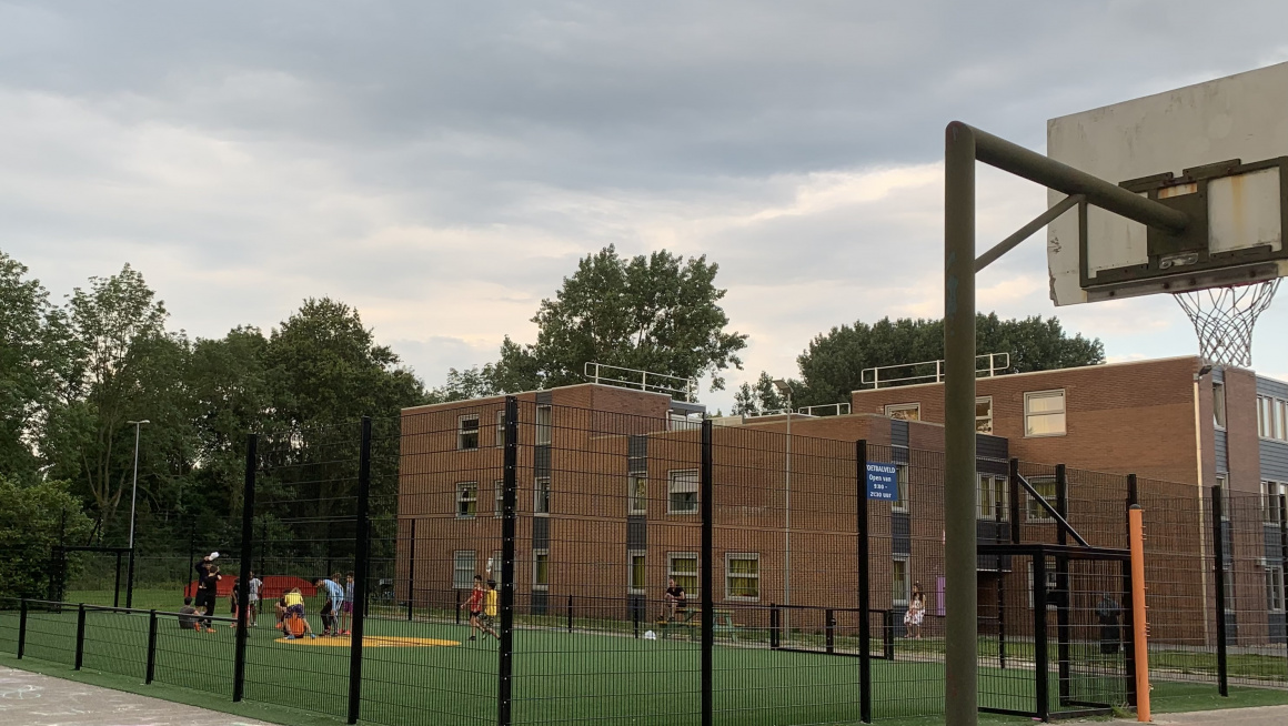 Cruyff court met op de voorgrond rechts een basket, daarachter een met hoge hekken omheind voetbal-/speelveld en daarachter woongebwouen met plat dak