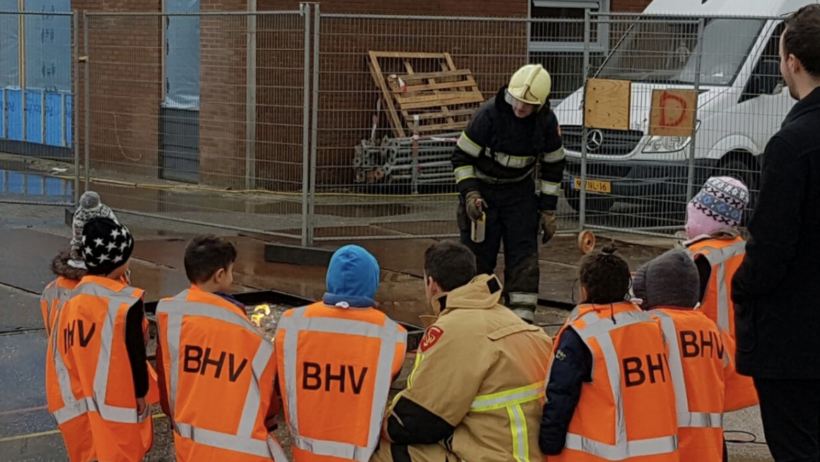 Kinderen met oranje hesjes met BHV erop met daartussen zittend op zijn hurken een brandweerman, allemaal kijkend naar een brandweermanmet helm die vuur maakt