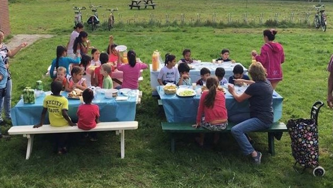 Een grasveld met daarop vier gedekte picknicktafels met kinderen en begeleiding die samen als kinderactiviteit eten