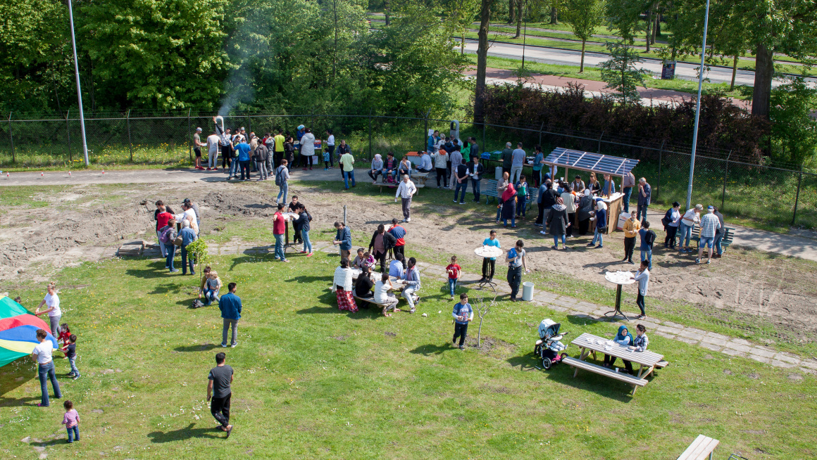 Zomerfeest op een grasveld met rondom bomen, op het grasveld picknick- en statafels, een kraampje en een barbecue met overal bewoners