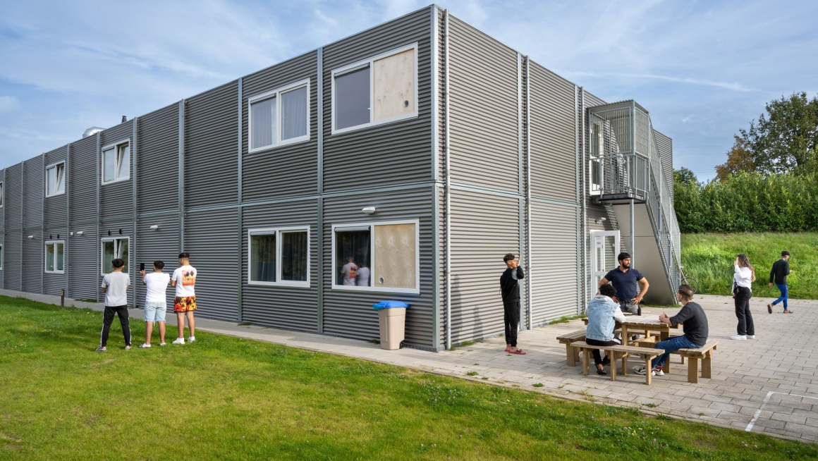 Foto van een woongebouw op een azc, met enkele bewoners op een grasveld en aan een picknicktafel