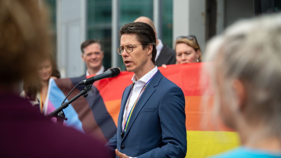 Bestuurslid, donker haar, donkerblauw pak staat voor gekleurde progressvlag