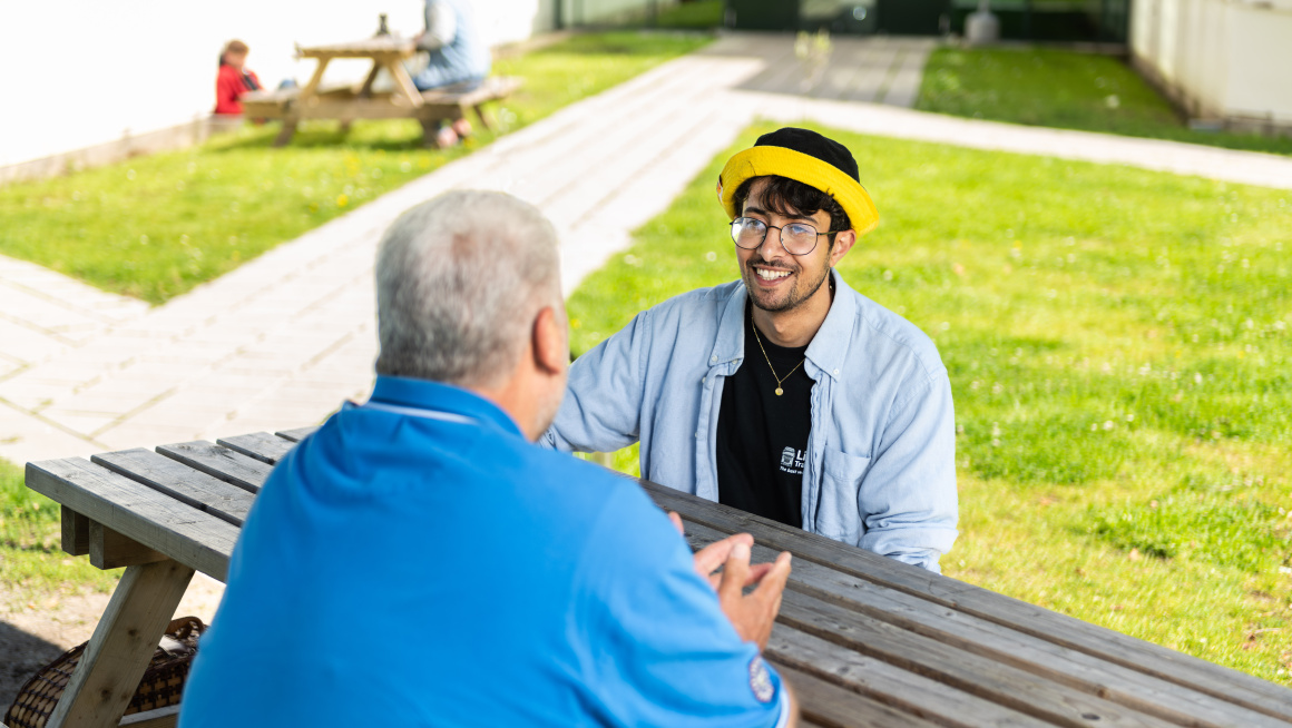 jongeman in gesprek met meneer buiten aan tafel in azc