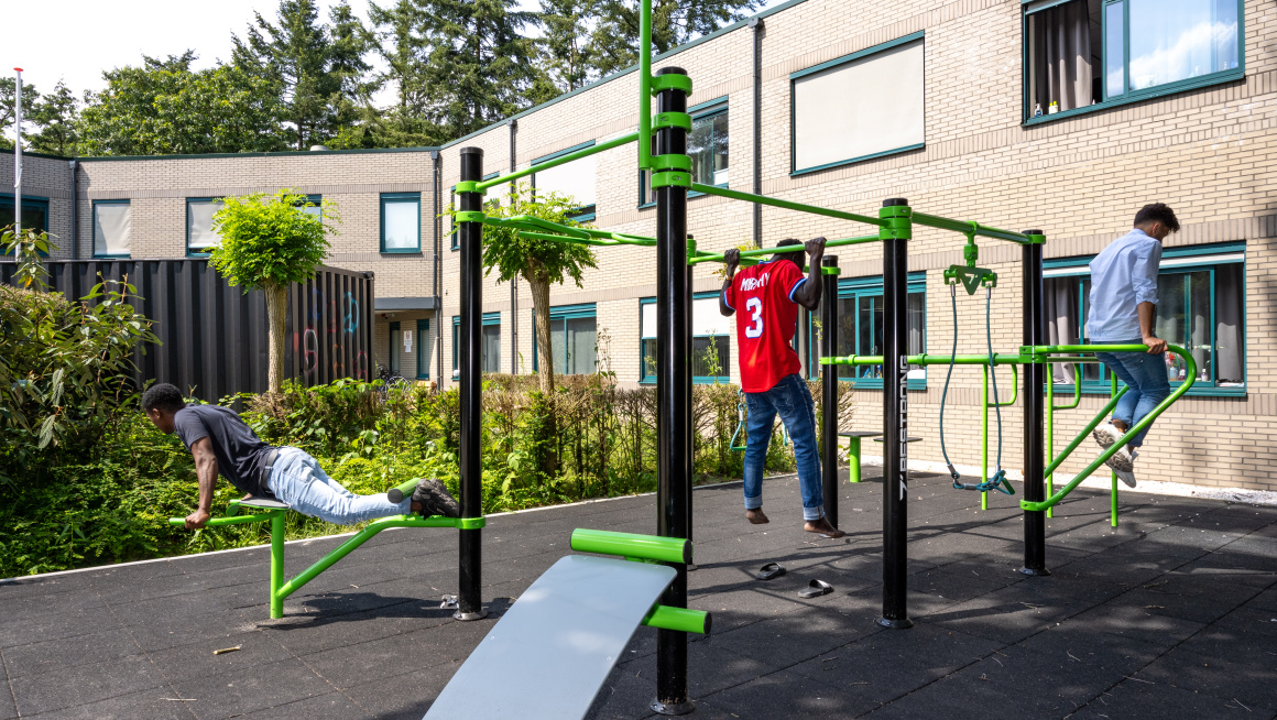 3 jonge mannelijke asielzoekers zijn buiten aan het sporten op groene sporttoestellen bij het azc Nunspeet. Ze doen opdrukoefeningen.