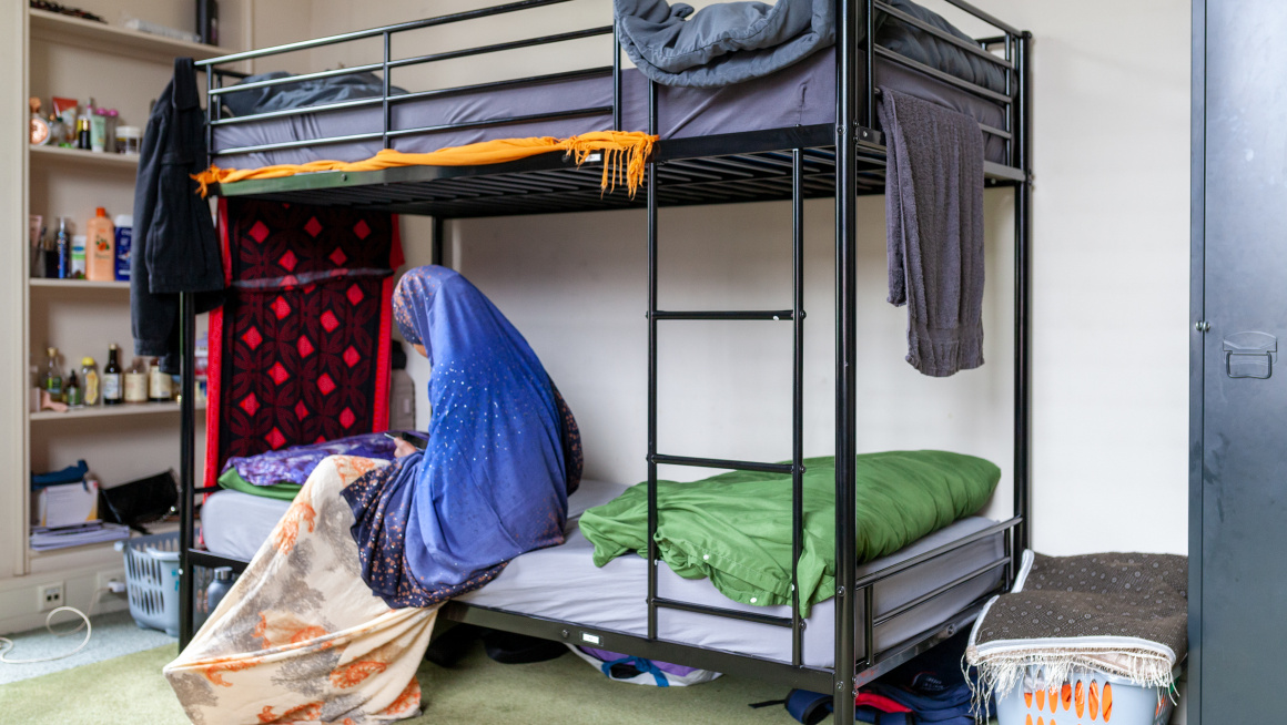 Een amv meisje zit in haar slaapkamer op het onderste bed van een stapelbed en kijkt op haar telefoon. Je ziet haar vanaf de linkerzijkant. Ze draagt een blauwe hijab en een lange jurk. De open kast links achter het bed staat vol met spulletjes waaronder shampoo. Er staat een volle wasmand rechts naast het bed. Aan het bed hangen handdoeken te drogen.