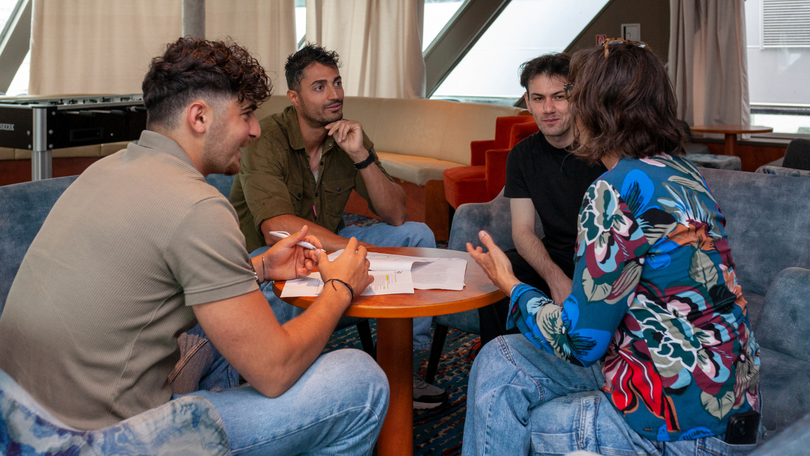 Drie jonge mannelijke statushouders zijn in gesprek met een vrouwelijke woonbegeleider in de salon van het schip waar zij verblijven. De woonbegeleider vertelt iets waar de mannen om moeten lachen. Ze zitten op blauwe banken rondom een ronde houten tafel. Er liggen papieren op tafel en de linkerman houdt een pen vast.