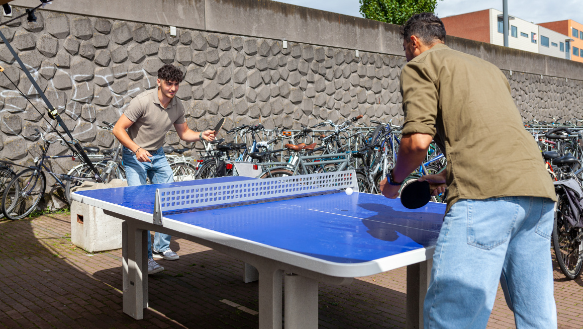 Twee jonge mannelijke statushouders zijn buiten op de kade voor het schip waar zij verblijven een potje aan het tafeltennissen. De zon schijnt. Er staan heel veel fietsen om hen heen geparkeerd.