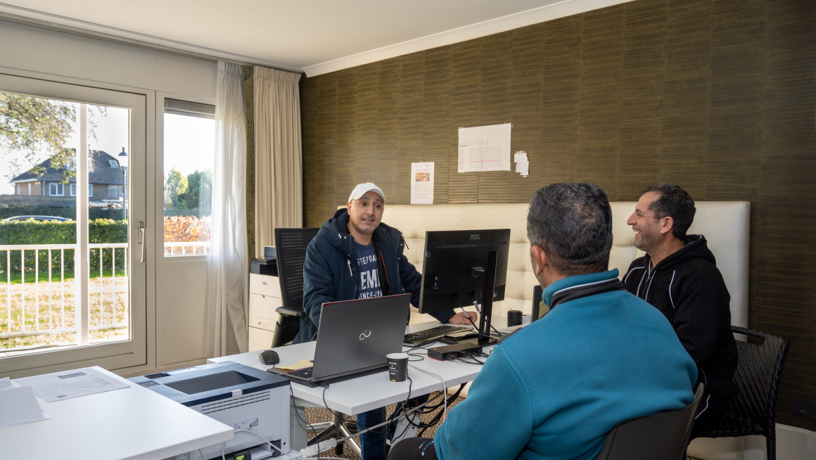 Twee asielzoekers in gesprek met een medewerker die achter de laptop zit.