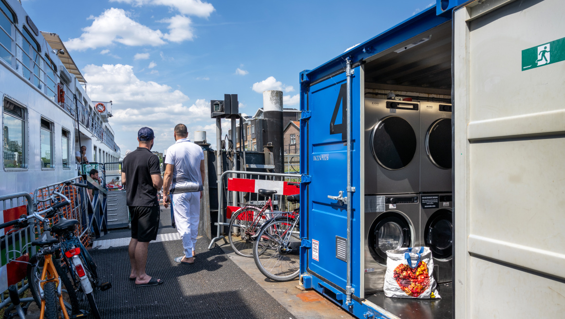 Twee mannen staan tussen een container met wasmachines en de asielboot in.