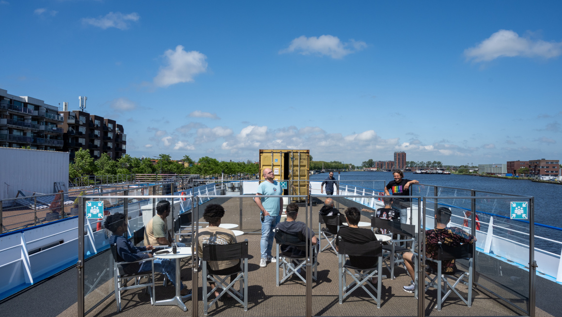 7 asielzoekers zitten op het dek van hun asielboot en luisteren naar twee medewerkers. Allen zijn mannen.