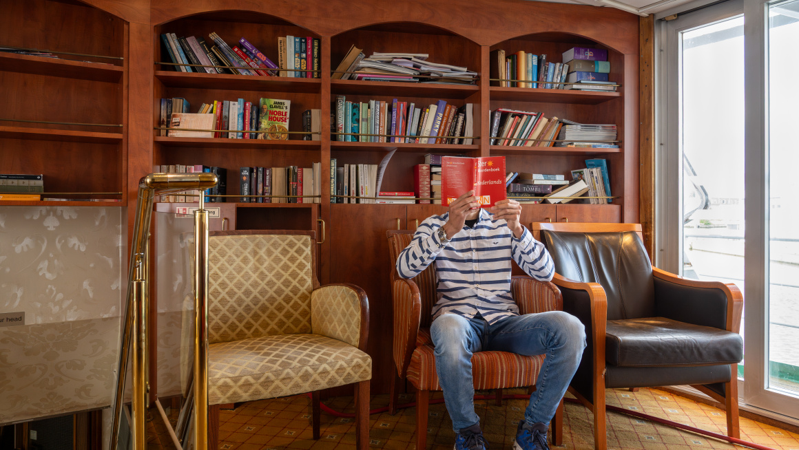 Man zit op een stoel die voor de boekenkast staat een boek te lezen.