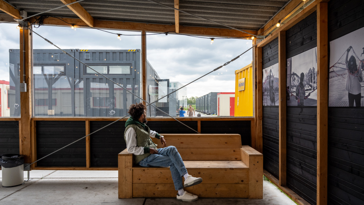 Een man zit op een houten bank onder een afdak en kijkt de straat met wooncontainers in.