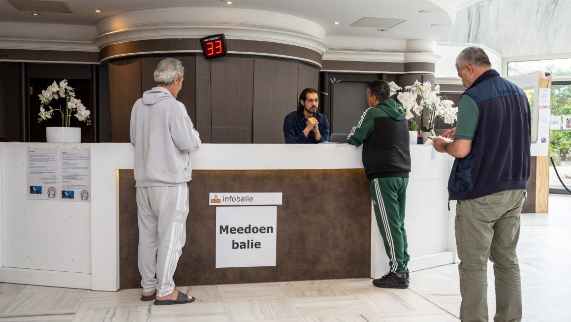 Drie mannen staan in de rij bij de Meedoenbalie, de medewerker is met één van hen in gesprek.