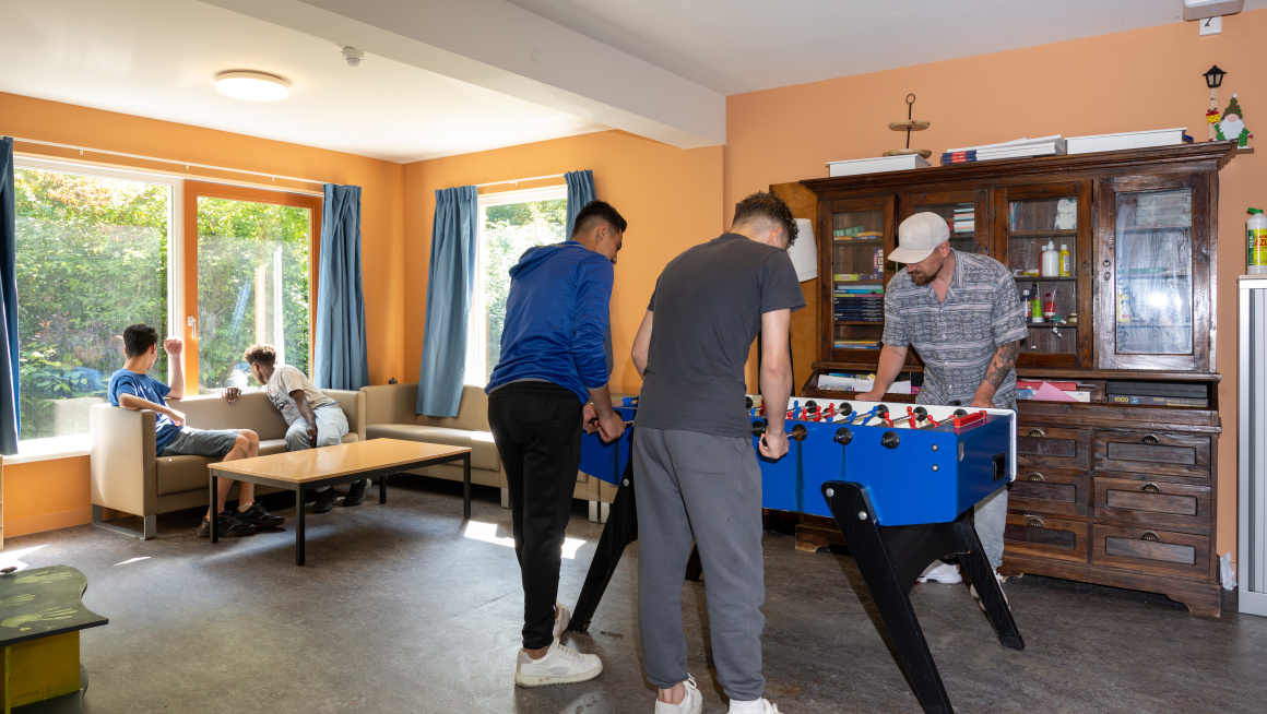 2 minderjarige jongens spelen tafelvoetbal met een medewerker. Op de bank zitten 2 jongens die naar buiten kijken.