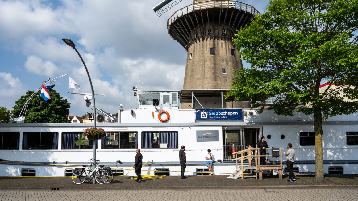 Asielzoekers buiten op de kade bij de ingang van de asielboot. Op de achtergrond staat een molen.