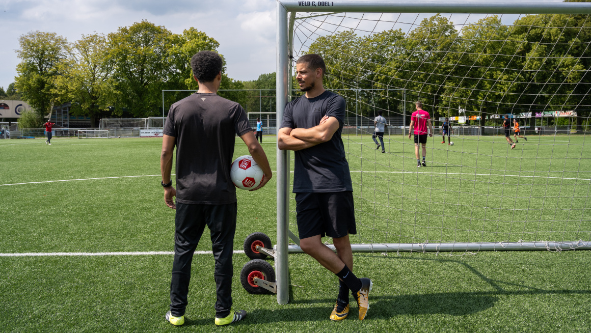 Keeper leunt tegen doelpaal en trainer heeft bal onder zijn arm