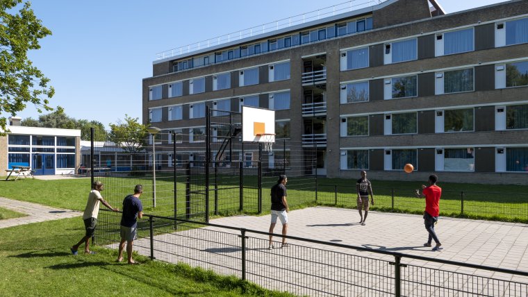 grasveld met betegeld basketbalveld met basketballende bewoners en op de achtergrond het woongebouw