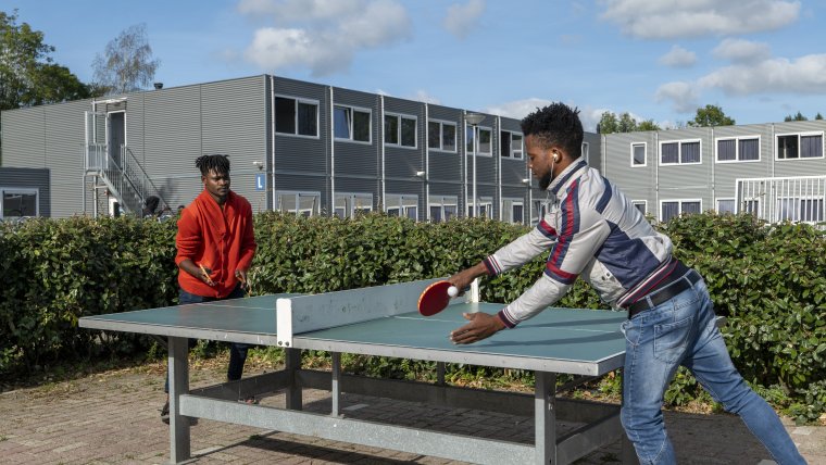 Bewoners spelen buiten tafeltennis voor struiken met daarachter grijze woongebouwen