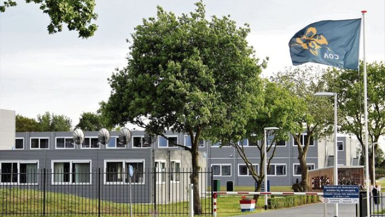 Voorkant azc met ingang met slagboom, COA vlag, grijze gebouwen en bomen