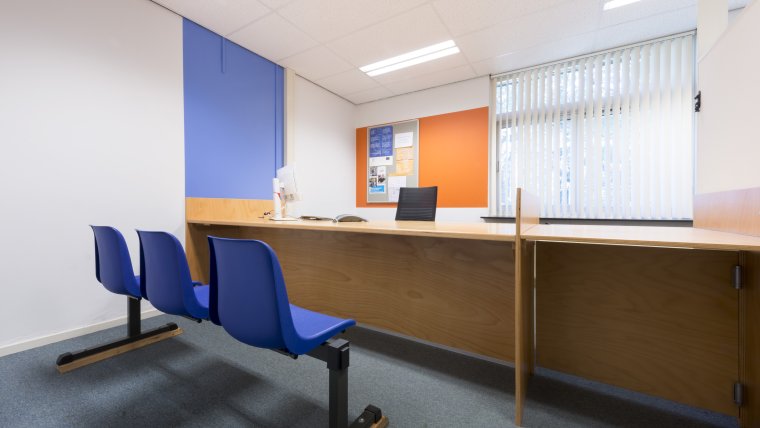 spreekkamer met op de voorgrond 3 blauwe stoelen, daarachter een bureau en daaracher een raam met witte lamellen, de muren zijn wit, blauw en oranje