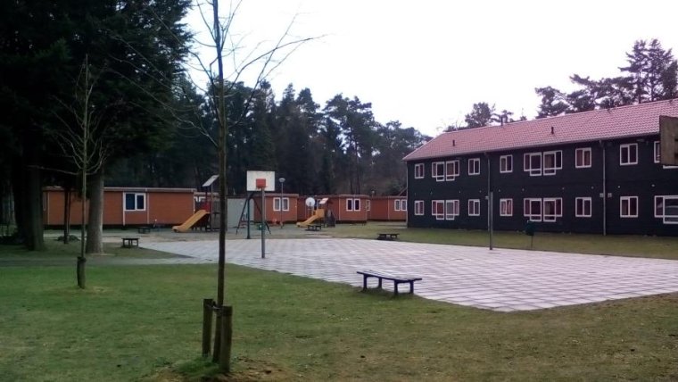 zwart gebouw met twee verdiepingen en rood dak met daarvoor gras en een betegeld basketbal-/volleybalveld en daarnaast 4 geel/bruine stacaravans met speeltoestellen ervoor