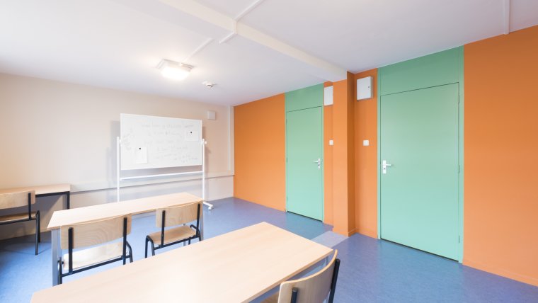Leslokaal met witte en oranje muren en mintgroen deuren, tafels en stoelen en een whiteboard