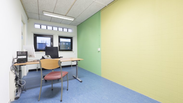 kantoorruimte met bureau, bureaustoel en gewone stoel ervoor. Blauwe vloer en witte, groene en gele muren en twee ramen