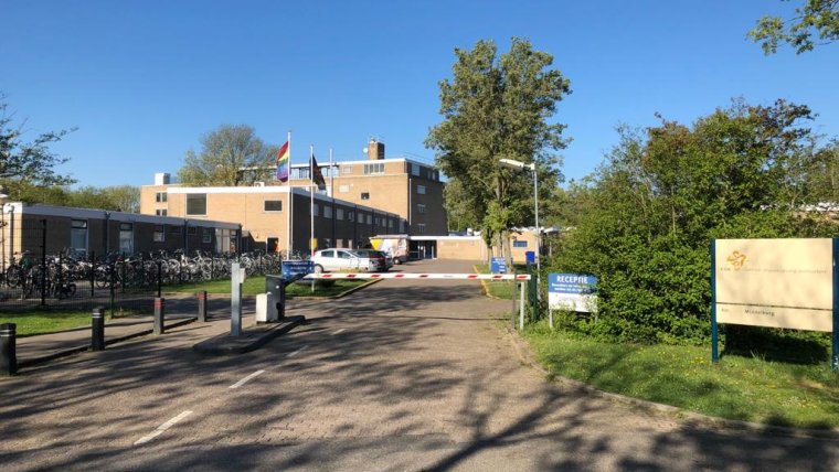 ingang azc met slagboom en COA bord met naam azc daarachter gebouwen, gras, struiken en bomen