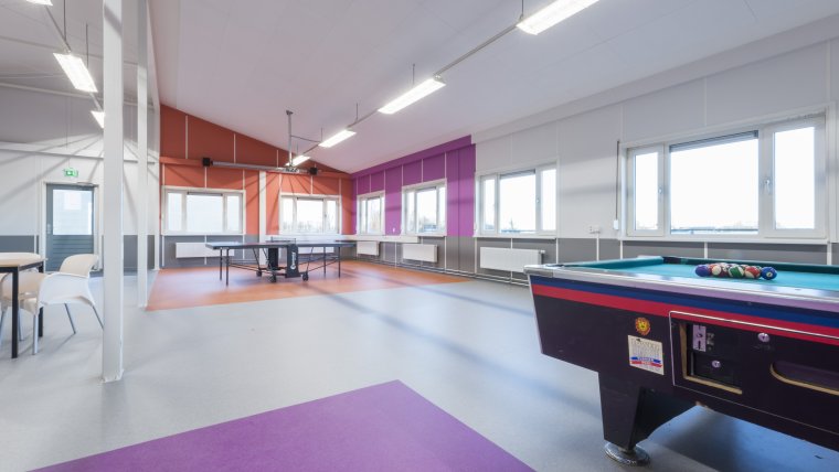 Recreatieruimte met grijze vloer met paarse en oranje vlakken en witte, paarse en oranje muren, een tafel met stoelen, een biljarttafel en een tennistafeltafel