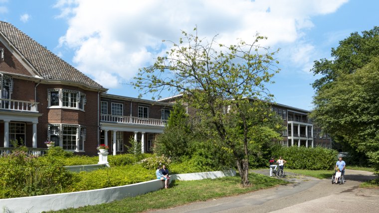 buitenkant kantoorgebouw met witte pilaren en witte balkons. Daarvoor struiken, bomen, wit laag muurtje en een pad met daarop wandelende bewoners