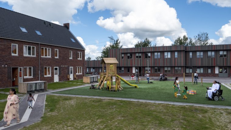 woongebouwen met daarvoor gras, speeltoestellen en paden met wandelende en spelende bewoners