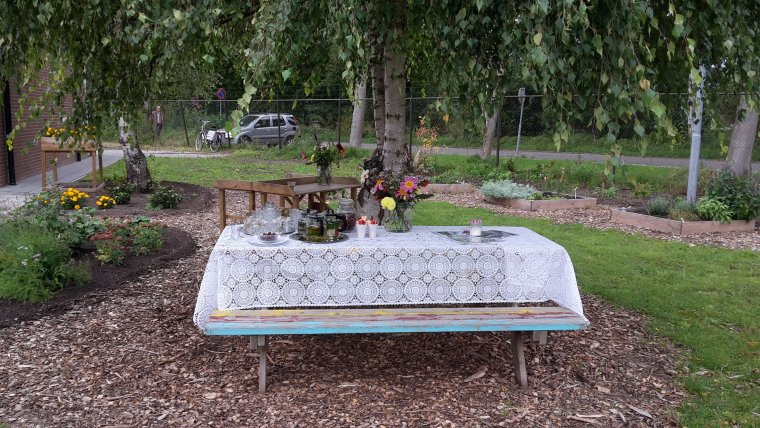 Picknicktafel met daarop een kleed en decoratie zoals bloemen onder de tafel liggen houtsnippers en daaromheen ligt gras en perkjes met struiken, bloemen en bomen