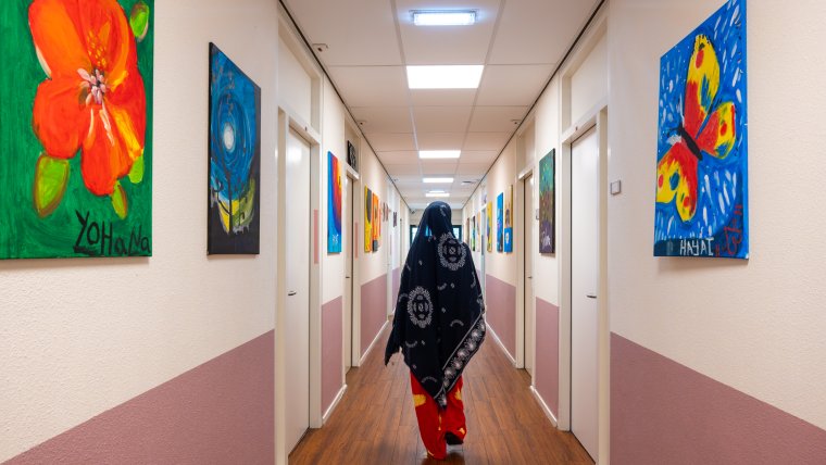 Jonge vrouwelijke asielzoeker in jurk loopt door de gang in het azc, we zien haar op de rug, er hangen kleurrijke schilderijen aan de muur
