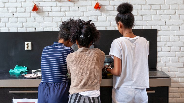 Drie amv meisjes doen de afwas aan een zwart aanrecht, erboven hangen rode kopjes aan de witte muur. We zien de meisjes op de rug. Ze hebben zwart, opgestoken haar. De linker draagt een blauw gestreepte trui, de middelste een beige trui en de rechter een wit t-shirt.