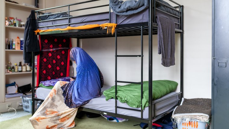 Een amv meisje zit in haar slaapkamer op het onderste bed van een stapelbed en kijkt op haar telefoon. Je ziet haar vanaf de linkerzijkant. Ze draagt een blauwe hijab en een lange jurk. De open kast links achter het bed staat vol met spulletjes waaronder shampoo. Er staat een volle wasmand rechts naast het bed. Aan het bed hangen handdoeken te drogen.