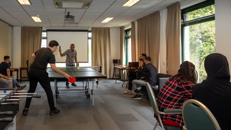 twee mannen spelen tafeltennis in de recreatieruimte waar meerdere mensen aan de kant zitten en kijken