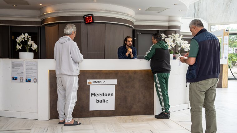 Drie mannen staan in de rij bij de Meedoenbalie, de medewerker is met één van hen in gesprek.