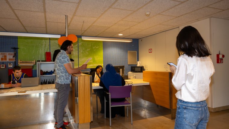 Een man en een vrouw wachten bij de infobalie. Een medewerkster is ingesprek met een vrouw die aan de balie zit.