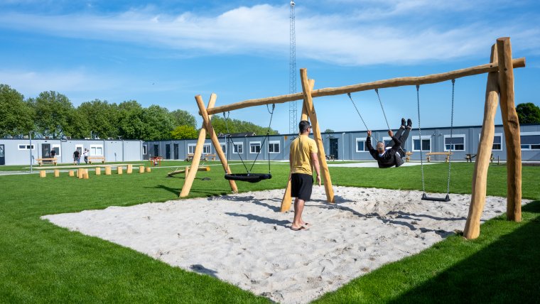 2 mannelijke asielzoekers op de schommel in de speeltuin naast de noodopvang.