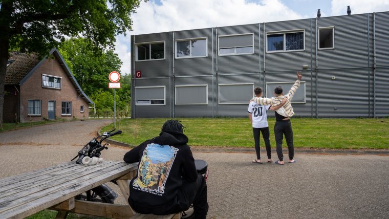3 jongens voor het gebouw, eentje zit aan een picknicktafel de andere staan met de armen om elkaar heen geslagen