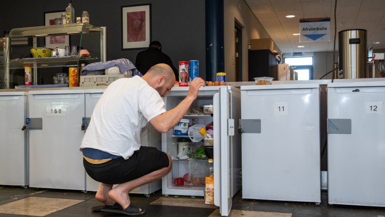 een man zit gehurkt voor zijn open koelkast, er staan links en rechts meer koelkasten met kamernummers erop